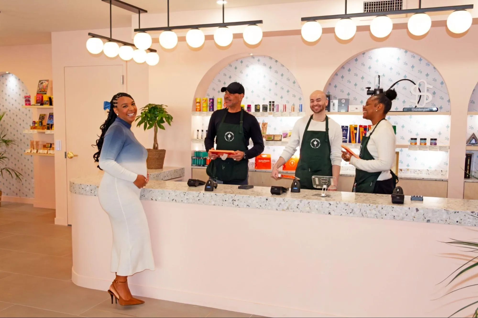 A customer smiling at the checkout counter while chatting with three friendly budtenders in green aprons at Yerba Buena