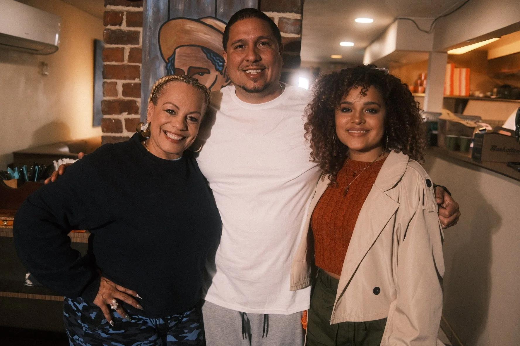 Three people smiling and standing together in a cozy indoor setting with brick wall