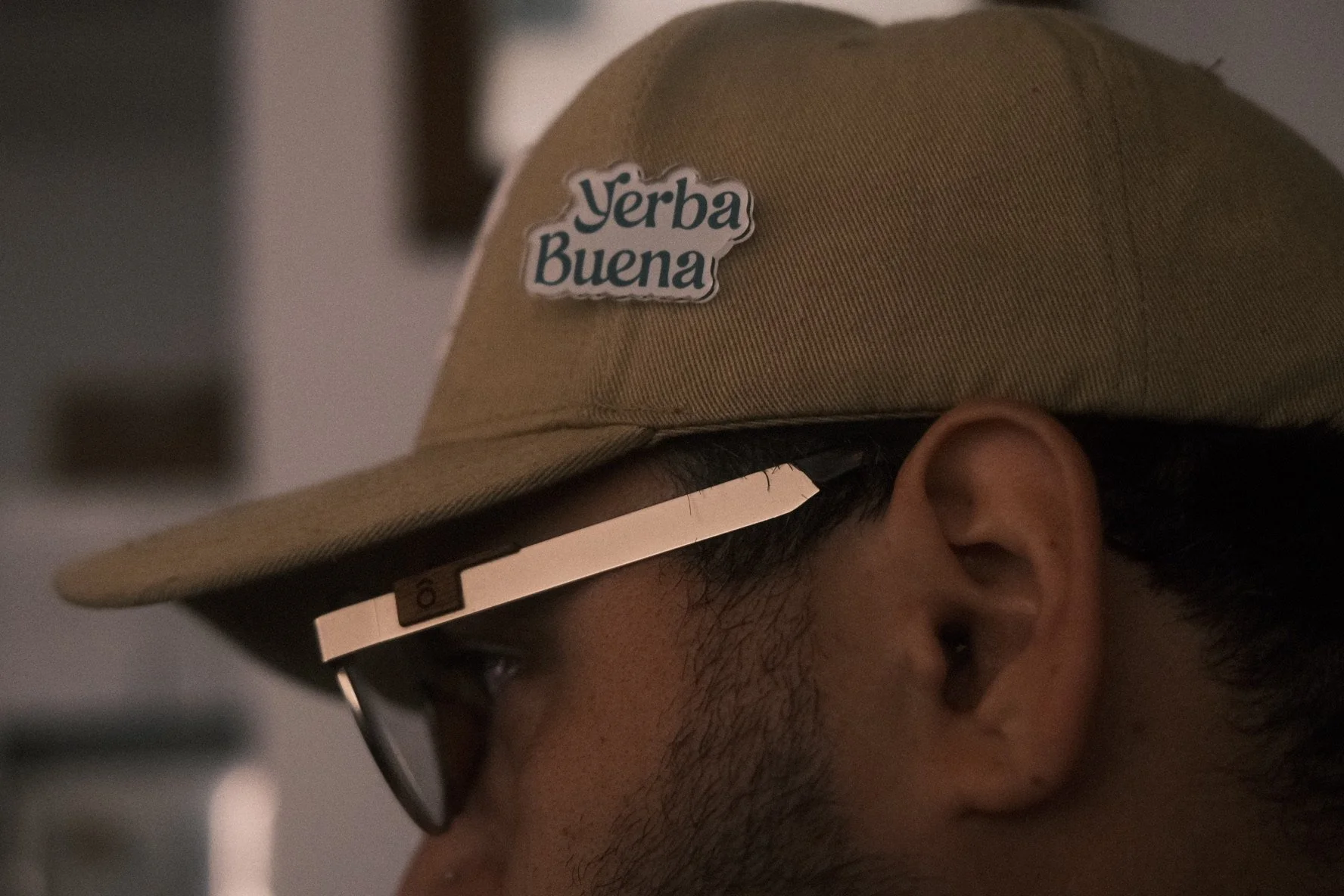 Close-up of a man wearing a tan cap with a Yerba Buena sticker and glasses