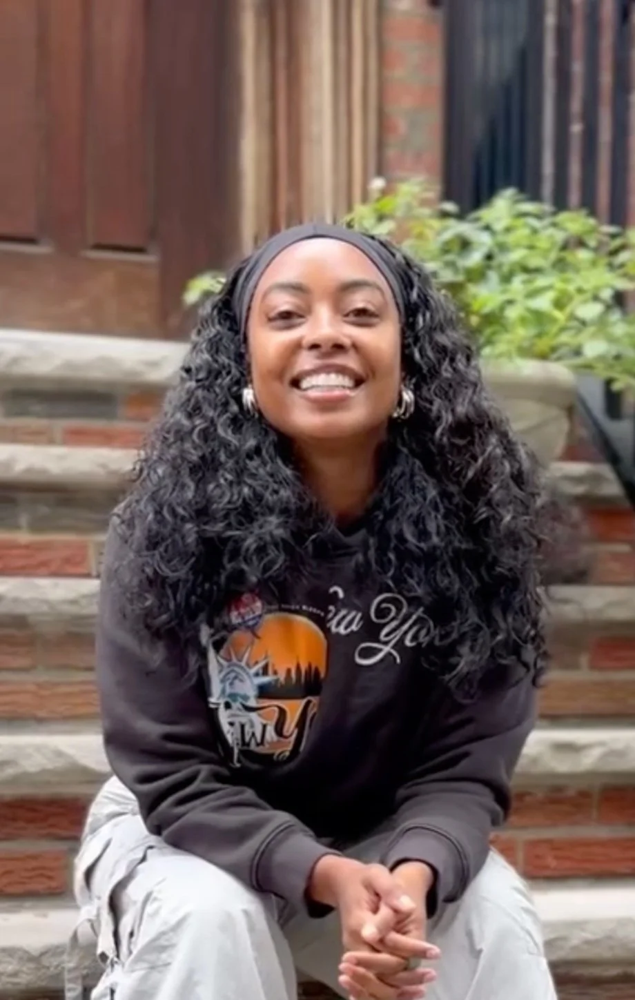 A woman with long, curly black hair wearing a gray hoodie sitting on outdoor stairs