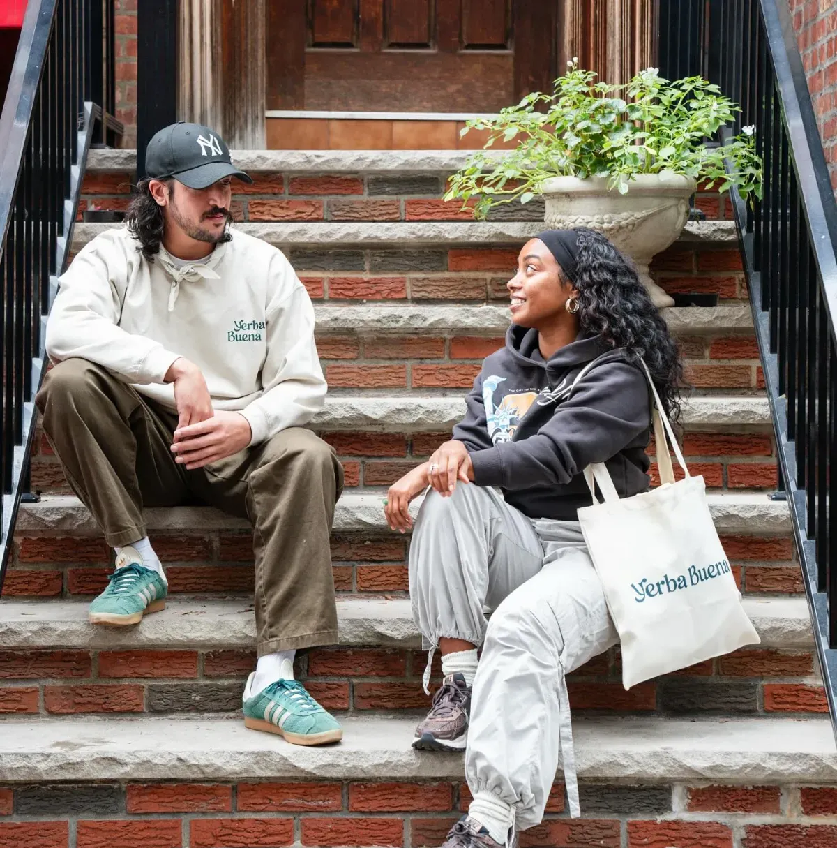 Two curious people wearing Yerba Buena merchandise smiling at each other while sitting on outdoor steps.